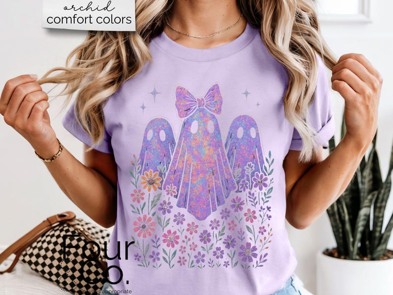Puede incluir: Camiseta lila claro con un dise&ntilde;o de tres fantasmas con un lazo, rodeados de flores. Los fantasmas tienen un efecto tie-dye colorido. El texto "orchid comfort colors" est&aacute; en la esquina superior izquierda.