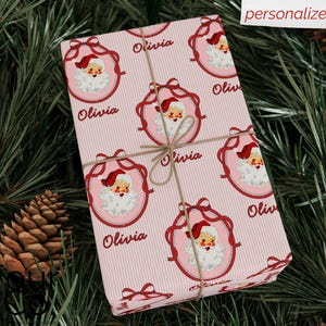 May include: A wrapped gift with pink striped paper featuring Santa Claus illustrations in red frames. The name "Olivia" is printed on the paper. The gift is tied with twine and rests on a bed of pine needles and pine cones.