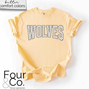 May include: A light yellow t-shirt with the word "WOLVES" in large, black, polka-dotted letters across the front. The shirt has rolled-up sleeves and the text "butter comfort colors" is in a white box in the upper left corner.