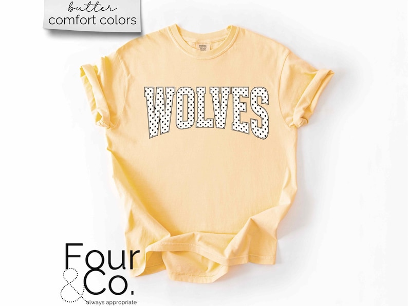 May include: A light yellow t-shirt with the word "WOLVES" in large, black, polka-dotted letters across the front. The shirt has rolled-up sleeves and the text "butter comfort colors" is in a white box in the upper left corner.