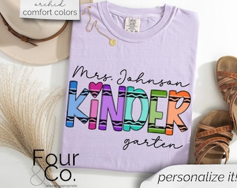 May include: A lavender t-shirt with the text "Mrs. Johnson Kindergarten" in colorful crayon-like lettering. The text is surrounded by a heart and a rainbow.