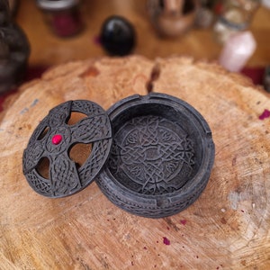 Pode incluir: Uma caixa de joias preta com um design de n&oacute; celta e um acento de joia vermelha na tampa. A caixa est&aacute; aberta e a tampa est&aacute; apoiada em uma superf&iacute;cie de madeira.