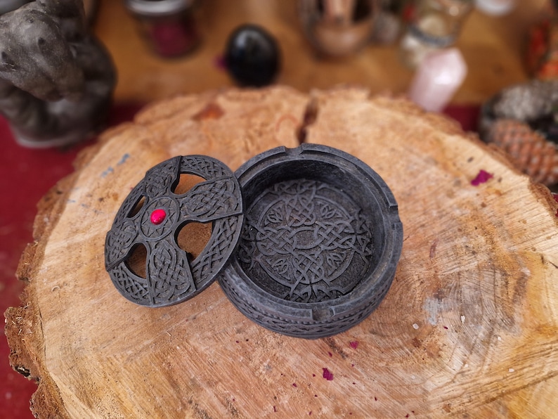 Pode incluir: Uma caixa de joias preta com um design de n&oacute; celta e um acento de joia vermelha na tampa. A caixa est&aacute; aberta e a tampa est&aacute; apoiada em uma superf&iacute;cie de madeira.