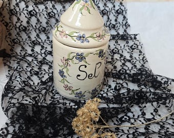 May include: A cream-colored ceramic canister with a conical lid, featuring hand-painted blue and pink floral vines and the word Sel written in black script on the front. The cylindrical container has a smooth finish and a matching floral pattern.