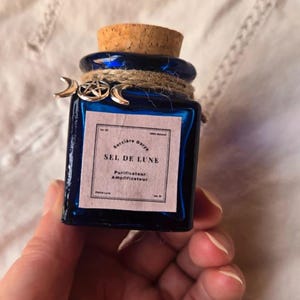 Peut inclure: Petite bouteille carrée en verre bleu foncé avec bouchon en liège. L'étiquette porte l'inscription "SEL DE LUNE" et des breloques décoratives argentées. La bouteille est entourée de ficelle.