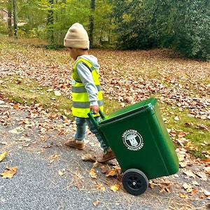 May include: A small green plastic rolling trash bin with a hinged lid and black wheels. The side of the bin features a white circular logo with the text Sanitation Department and a central graphic of a trash can icon with a handle.