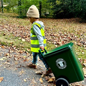 Op de afbeelding: Een kind met een beige gebreide muts, een grijze hoodie, een blauwe spijkerbroek en een felgeel veiligheidsvest met reflecterende strepen, dat een donkergroene plastic rolcontainer trekt met het opschrift City of Chamblee Sanitation Department.