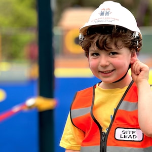 May include: A child wearing a white hard hat with the text SCHWARTZ BROTHERS CONSTRUCTION and an orange high-visibility safety vest with reflective silver stripes and a white rectangular patch featuring the black text SITE LEAD on the front.
