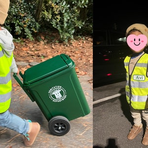 May include: A child dressed in a yellow safety vest and tan beanie pulls a green toy trash can with black wheels. The trash can has a white Sanitation Department logo. The child is wearing blue jeans and brown shoes.