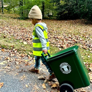 May include: A child wearing a beige knit beanie, a gray hooded sweatshirt, blue jeans, and a bright yellow high-visibility safety vest with reflective stripes, pulling a dark green plastic rolling trash bin labeled City of Chamblee Sanitation Department.