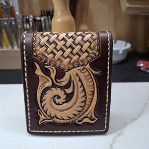 May include: Hand-tooled leather wallet with a basket weave pattern on the top flap and a detailed floral design on the front. The wallet is brown with tan accents and white stitching.