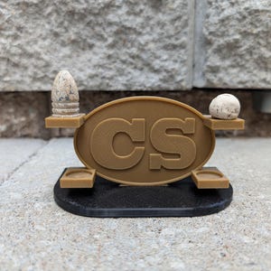 May include: A brown and black decorative display featuring the letters "CS" in a gold oval. The display has two small shelves holding a bullet and a round stone. The base is black.