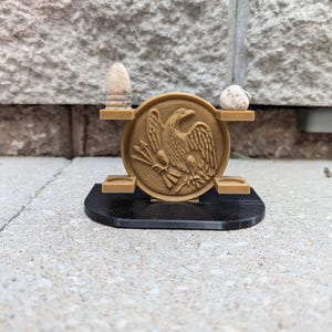 May include: A tan 3D-printed display stand featuring a circular emblem with an eagle design. Two small shelves hold a bullet and a rock. The stand has a black base and is set against a textured stone wall.