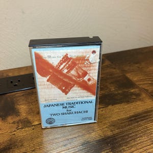 May include: A cassette tape case featuring a cover with a red and white graphic of a shakuhachi flute. The text reads Japanese Music for Two Shakuhachi, Lyrichord LLCT 7386, and includes artist names. The case has a black plastic top edge.