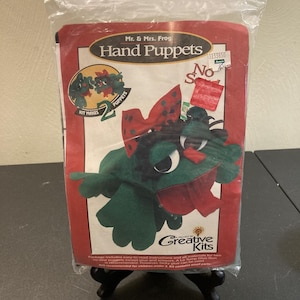 May include: A packaged hand puppet kit featuring a green frog puppet with a red bow and a red border. The package reads "Mr. & Mrs. Frog Hand Puppets" and "Creative Kits". The kit makes two puppets.