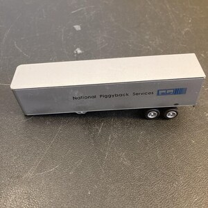 May include: A silver-colored toy semi-trailer truck with the text National Piggyback Services printed in black on the side, accompanied by a blue logo. The trailer features a rectangular body and is supported by two black wheels on the rear axle.