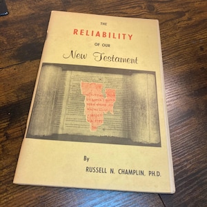 May include: A softcover book titled The Reliability of Our New Testament by Russell N. Champlin, Ph.D. The cover is light tan with red and black text and features a central illustration of an ancient scroll with a red fragment overlay.