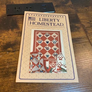 May include: A rectangular paper pattern booklet titled Liberty Homestead featuring a quilt design with red and white striped fabric, basket motifs, and small rabbit figures. The cover displays a central quilt image with a blue border and stars.