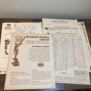 May include: A collection of white paper instruction manuals and charts for the MEC 600 Jr. shotshell reloader. The documents feature black printed text, technical diagrams, and tables detailing charging bars, wad pressure, and powder bushing specifications.