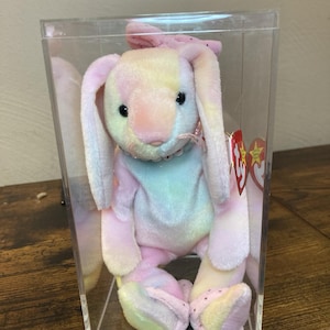 May include: A plush rabbit toy with pastel tie-dye fur in shades of pink, yellow, and blue, enclosed in a clear rectangular display case. The rabbit features long ears, black eyes, a beaded collar, and a red Ty heart-shaped tag attached to its side.