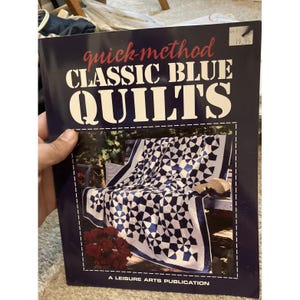 May include: A softcover instructional book titled Quick-Method Classic Blue Quilts, a Leisure Arts Publication. The dark blue cover features a photograph of a white and blue patterned quilt with a circular design, displayed on a wooden bench.