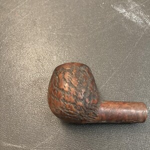 Puede incluir: Una pipa de tabaco marrón con un cuenco texturizado y una boquilla recta. La pipa es de madera y tiene un color oscuro y rico. El cuenco es redondeado y la boquilla es cilíndrica.