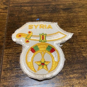 May include: An embroidered patch with a white background featuring the word SYRIA in yellow letters. The design includes a yellow and white curved sword above a yellow crescent moon and star emblem with red and green accents on the edges.