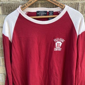 May include: A long-sleeve raglan shirt with a deep red body and cream-colored sleeves. The chest features a white graphic logo reading Polo Jeans Company with an RL emblem. The interior neck label displays Ralph Lauren Polo Jeans Co. text.