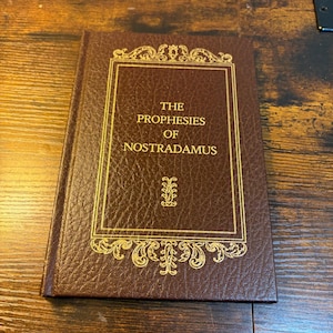 May include: A brown hardcover book titled The Prophesies of Nostradamus, featuring a textured cover with gold foil lettering and ornate decorative borders surrounding the text and a small central emblem on the front cover.