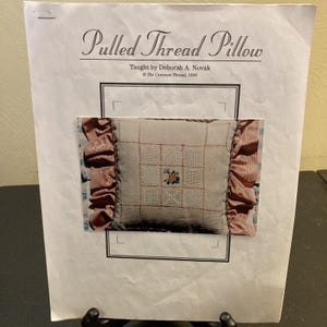 May include: A printed paper titled "Pulled Thread Pillow" with a picture of a decorative pillow. The pillow has a light-colored center with a floral design and a ruffled border. The text on the paper includes the name of the teacher and the copyright information.