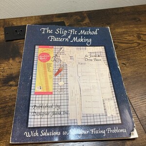 May include: A softcover instructional book titled The Slip-Fit Method of Pattern Making for Jeans & Dress Pants, published by Designer Jeans, Inc. The dark blue cover features a grid background, sewing pattern pieces, and a yellow fashion ruler.