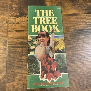 May include: A vertical rectangular booklet titled THE TREE BOOK with a green background. The cover features a photograph of a person holding a small tree wrapped in newspaper, surrounded by images of trees. Text includes $2.00 and FREE RED MAPLE TREE.