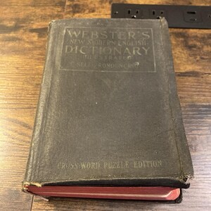 May include: A black hardcover Webster's New Modern English Dictionary Illustrated Self-Pronouncing Cross Word Puzzle Edition book with visible wear on the edges and a red-tinted page edge visible at the bottom of the thick volume.