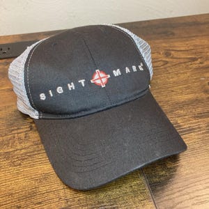 May include: A black baseball cap with a gray mesh back and a curved brim. The front panel features the embroidered white text SIGHT MARK with a red and white crosshair symbol replacing the letter A. The cap has a structured crown and stitching.