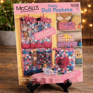May include: A McCall's sewing pattern envelope titled Pretty Doll Pockets, pattern number 15218. The cover features a pink and blue checkered fabric organizer with pockets for dolls, a matching tote bag, and text stating it holds 8 dolls plus accessories.