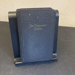 May include: A small, black, textured-cover book titled New Testament Psalms in gold lettering, resting upright in a black plastic display stand. The book features a pebbled leather-like finish and rounded corners, held securely by the stand's frame.