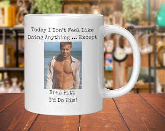 May include: White ceramic mug with a handle, featuring a photo of Brad Pitt and the text "Today I Don't Feel Like Doing Anything... Except Brad Pitt I'd Do Him!" The mug is on a wooden surface.