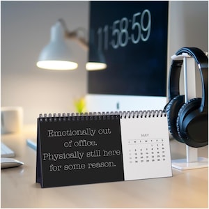 以下が含まれることがあります： 「Emotionally out of office. Physically still here for some reason.」と書かれた黒い表紙の卓上カレンダー。カレンダーには5月が表示されています。黒いヘッドフォンが白いスタンドに置かれています。