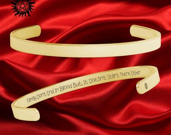 May include: Gold-colored cuff bracelet with the engraved text "Family Don't End In Blood But, It Doesn't Start There Either." The bracelet has rounded edges and a smooth finish. A black and red symbol is in the upper left corner.