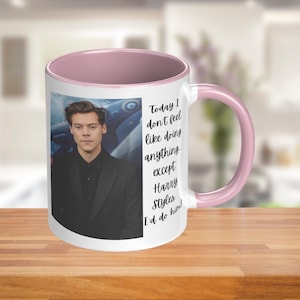 Puede incluir: Una taza de cerámica blanca con un borde y un asa de color rosa. La taza presenta una foto de un hombre con cabello oscuro y una chaqueta negra. El texto en la taza dice "Today I don't feel like doing anything except Harry Styles. I'd do him!"