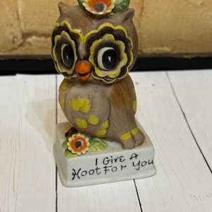 May include: A ceramic owl figurine with a floral crown and yellow accents. The owl has large eyes and is perched on a rectangular base with the text "I Give A Hoot For You". The figurine is brown, yellow, and orange.
