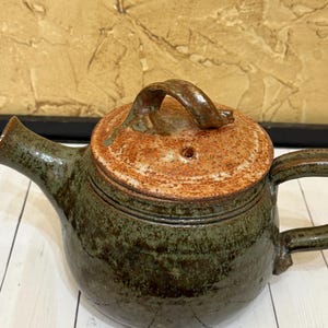 May include: Handmade ceramic teapot with a dark green body and a reddish-brown lid. The teapot has a curved spout and a handle on the side. The lid has a small handle and a hole for steam. The teapot is sitting on a white wooden surface.