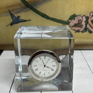 May include: A clear, square quartz clock with a silver-colored face and hands. The clock face has black Roman numerals and minute markers. The clock is encased in a transparent, cube-shaped block.
