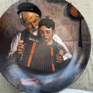 May include: Decorative plate featuring a painting of an older man and a young boy playing an accordion. The man wears a black beret and glasses, while the boy has dark hair. The plate has a dark border and the artist's name, Norman Rockwell, is printed on the plate.