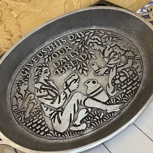 May include: Oval, pewter-colored serving dish with a detailed relief design. The design features figures, a basket, and the text "GIVE US THIS DAY OUR DAILY BREAD". The dish has a slightly raised rim and a handle on each side.