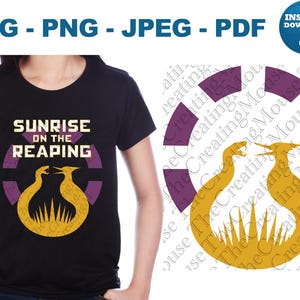 Könnte beinhalten: Ein schwarzes T-Shirt mit einem Grafikdesign und dem Text SUNRISE ON THE REAPING in weißen Buchstaben. Unter dem Text befindet sich ein kreisförmiges violettes Emblem mit zwei gelben Vögeln, die sich über einem Sonnenstrahlenmuster gegenüberstehen.