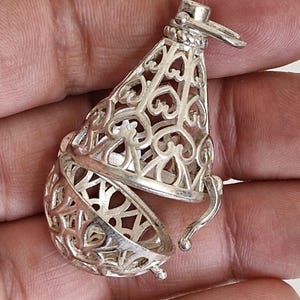 May include: A silver-colored metal pendant featuring an intricate, openwork filigree design with repeating heart-shaped motifs. The pendant has a conical top section and a hinged, spherical bottom portion that opens to reveal a hollow interior space.