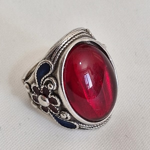 May include: A silver ring featuring a large, oval, translucent red cabochon stone. The metal band is decorated with intricate silver wirework and small floral motifs accented with dark blue enamel, creating a detailed and textured appearance.