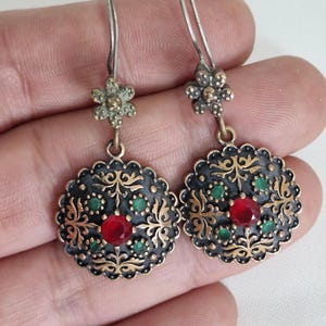 May include: A pair of drop earrings featuring a circular medallion with a scalloped edge, finished in an oxidized silver tone with gold-colored filigree accents. Each earring is set with a central red stone surrounded by four small green stones.