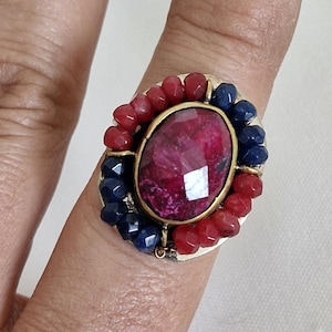 May include: A ring featuring a large, oval, faceted magenta center stone set in a gold-toned bezel. The center stone is surrounded by a border of alternating small, faceted red and dark blue beads, creating a vibrant, multi-colored circular design.